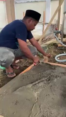 A man wearing a dark blue t-shirt, gray pants, and sandals is kneeling on the ground, smoothing out wet cement with a red-handled trowel. He also has a black cap on his head. The cement is contained within a wooden rectangular frame constructed from planks. A white tank and some hoses are visible to the right. The ground around the frame is a mixture of dirt and sparse grass. A bucket sits in the background. The setting appears to be an outdoor or semi-enclosed space, possibly a backyard or under an open-sided structure.