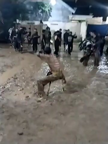 The video captures an outdoor nighttime scene in Jenggrik, Indonesia, featuring a group of people, primarily young men, engaged in an activity in a deeply muddy area. The ground is heavily saturated with mud, suggesting recent rain or intentional mudding for the event. Artificial lights illuminate the scene, with a white building visible in the background.

In the foreground, a person is prominently seen at 0:00 covered head-to-toe in mud, performing a spinning or dancing movement. They appear to be wearing a striped shirt and trousers, both caked in mud. At 0:01, this individual turns and walks away, their mud-splattered back visible.

Other participants are scattered throughout the muddy ground. Some are also covered in mud, while others wear dark clothing. There are individuals in costumes, including what appears to be a black and white horse-like costume (possibly for Jaranan or Kuda Lumping, a traditional Javanese dance) and other dark, mask-like outfits. The participants are moving around, some watching, others seemingly involved in the performance. The atmosphere appears to be lively and energetic, characteristic of a cultural performance or ritual. No discernible text is visible in the video.
