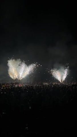 The video shows a large crowd of people silhouetted against a dark background, observing a fireworks display. Two plumes of white and yellow fireworks are erupting in the sky, creating a spray of sparks and smoke. The crowd appears to be densely packed, with many individuals holding up their phones, presumably to record the event. The overall atmosphere seems to be one of excitement and celebration.