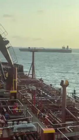The view is of the open sea, from the deck of a large oil tanker, facing forward. Another, similar sized tanker can be seen in the distance, a few miles away. The surface of the tanker in the foreground is crowded with piping, metal walkways, and other engineering structures. The water is blue with small waves, and the sky is overcast with a warm glow from the low-hanging sun.