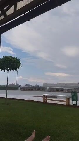 The video shows an outdoor scene with a person's feet visible in the lower left corner. In the mid-ground, there is a wooden bench and a green trash can next to a paved area. Beyond that, there is a body of water with fountains, and in the background, there are buildings and industrial structures under a cloudy sky. The camera pans to the right, revealing a shower fixture near the water's edge. The overall atmosphere appears to be overcast and possibly humid.