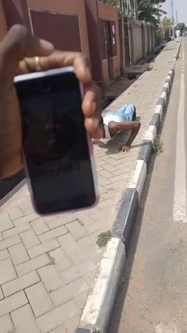 The video shows a person filming another person doing push-ups on a sidewalk next to a road. The sidewalk is made of rectangular light grey bricks, and is bordered by a curb painted with alternating black and white stripes. The person doing push-ups is wearing a white tank top and light blue shorts. They are facing the camera and are in the middle of performing a push-up. In the background, there is a brown building with a wired fence on top. There are also trees and a blue sky visible. A white vehicle passes by on the road during the recording.