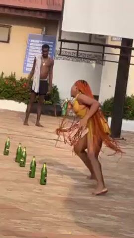 A woman in a short yellow outfit with long orange hair runs and kicks over a row of green beer bottles. A shirtless man with a towel around his neck stands watching in the background. There is a yellow beer crate and a sign on a building in the background. The woman kicks a bottle over and then picks one up and drinks from it.