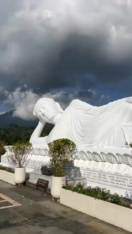 A low-angle, medium shot captures a massive, white reclining Buddha statue outdoors. The statue, which dominates the frame, is pristine white and detailed, with the Buddha resting on its side with its head propped on its arm. The statue is positioned on a large white platform with intricate carvings around its base. In front of the platform, a walkway with decorative paving stones is visible. Two large, white planters with small, leafy trees are placed to the left and right of the statue's base, and a low flower bed with purple flowers runs along the edge of the walkway in front of the statue. Behind the statue, lush green hills and mountains can be seen, topped with a dramatic sky filled with dark, heavy clouds interspersed with patches of bright blue sky and sunlight. The overall lighting suggests it is daytime.