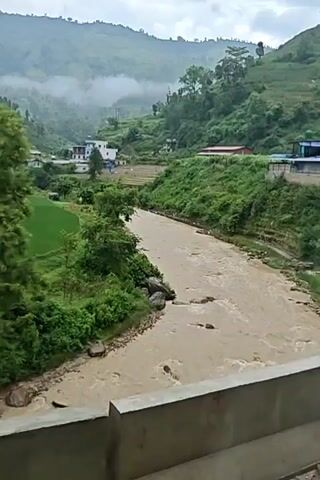 The video shows a river flowing through a valley with lush green hills. The river is brown and appears to be carrying a significant amount of sediment, suggesting recent rainfall. The banks of the river are lined with trees and vegetation. In the distance, there are a few buildings that appear to be houses or small structures, nestled amongst the greenery. The sky is overcast, with some clouds or mist hanging low in the hills, contributing to a serene and somewhat dramatic atmosphere. There are no people visible in the video. The overall impression is one of a natural landscape in a mountainous region.