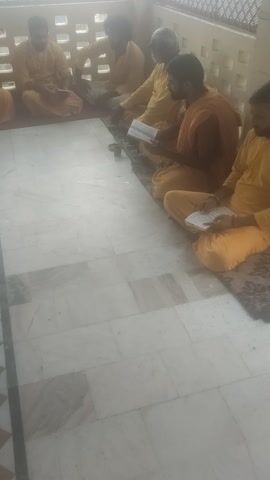 The video shows a group of six men, dressed in orange robes, sitting on a patterned rug on a tiled floor. They are arranged in a line, facing a wall with a decorative pattern of squares. Each man has a small white booklet in front of him, and they appear to be reading or reciting from them. Their hands are in a prayer-like position, with palms together. The lighting suggests it is daytime, with light coming from the left side of the frame. The overall atmosphere appears to be one of religious devotion or meditation.