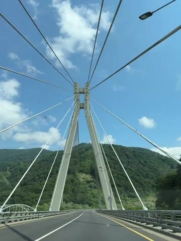 The video shows a view from a car driving across a cable-stayed bridge. The camera is looking forward through the windshield. The bridge has two tall, angled concrete towers that support numerous cables running to the road deck. The road is paved and has white lane markings. On either side of the road are metal railings. Beyond the bridge, lush green, tree-covered mountains rise under a blue sky with scattered white clouds. A few streetlights are visible along the bridge. There are no people or other vehicles clearly visible in the frame. The lighting suggests it is daytime.