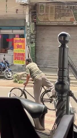 A man in a striped t-shirt and light-colored pants rides a bicycle down a street. His right leg is extended backward. A motorcycle is parked to his left. A sign for "Ashok Press" is visible behind him. The street has shops and a closed metal roll-up door on the right. A black structure with a spherical top is in the foreground on the right. The scene appears to be in India, judging by the signage.