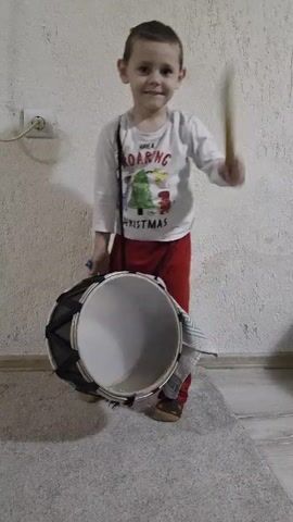 A young boy smiles and stands in front of a white wall with a drum hanging from his shoulder. The boy wears a white long-sleeve shirt with a "ROARING CHRISTMAS" graphic, red pants, and brown shoes. He holds a drumstick in his right hand and taps the drum. A wall outlet and wire are visible behind the boy. The floor is gray and white tile with a small gray rug behind him.