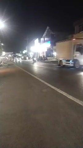 A video taken at night shows a street with businesses and traffic. Several cars and motorcycles are visible on the road, with their headlights illuminating the scene. The street appears wet, possibly from recent rain. Buildings line the right side of the street, with some of them lit up by interior lights and illuminated signs. A street lamp on the left side of the image provides additional lighting. The overall atmosphere is that of a busy urban or suburban street at night.