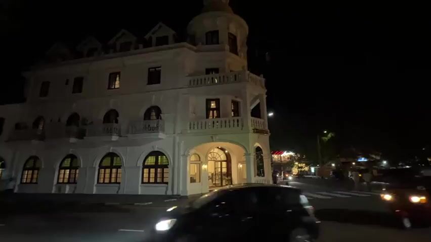 The video shows a street scene at night in front of a large white building.  The building has multiple levels, arched windows, and a rounded tower. It is lit up with interior lights.  In the foreground, cars and a tuk-tuk are driving past.  People are visible walking on a crosswalk and on the sidewalks. Streetlights illuminate the area, and trees are visible in the background.  The overall impression is a lively, but dimly lit, urban scene.