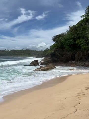 The video depicts a beach scene on a partly cloudy day. The beach has light-colored sand and the ocean waves are rolling in. Rocks are scattered along the shoreline, and there's a rocky cliff covered with green vegetation to the right. In the background, more lush greenery can be seen. A few footprints are visible in the sand. The sky has a mix of blue and white clouds.