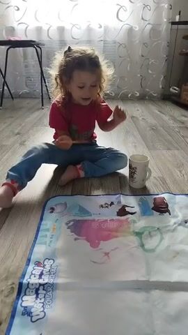 The video shows a young girl sitting on a wooden floor, engaged in an activity with a water-reveal coloring mat. The mat, laid out on the floor, features images of arctic animals, with some areas already colored in with vibrant hues. The girl, wearing a pink t-shirt and blue jeans, is holding a paintbrush, dipping it into a white mug filled with water, and then applying it to the mat. As she paints with water, colors magically appear on the mat, revealing the hidden details of the arctic landscape and its inhabitants.

The room appears to be a living space, with a window covered by sheer curtains in the background, suggesting it's daytime. Sunlight streams through, illuminating the scene and casting a soft glow. A stool and some shelves are visible in the background, hinting at a home environment. The girl's focused expression and the way she's carefully manipulating the brush indicate concentration and enjoyment in her artistic endeavor. The activity appears to be a form of creative play, likely providing a mess-free and engaging experience for the child. The mat itself has visible text that includes "Polar Animals" and "Find the hidden animals in the picture," guiding the child's play. The overall atmosphere is one of quiet, focused creativity within a home setting.