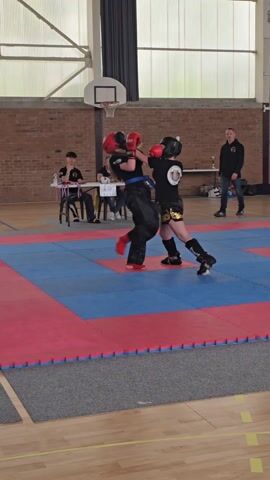 Two young women in full protective gear are engaged in a kickboxing match on a blue and red matted floor in what appears to be a gymnasium. The girl on the left is wearing black gear with gold accents, while her opponent on the right is wearing black gear with red gloves and helmet. They are actively punching and kicking, moving around the mat. In the background, a basketball hoop hangs from the ceiling, and several adults are watching the match, including some seated at a table with a timing device. The walls are made of brick and corrugated metal.
