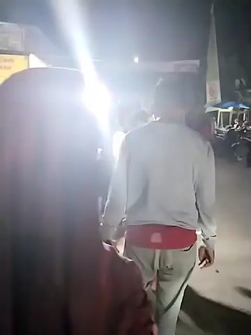 The video shows a night scene of a food stall. The stall is brightly lit and decorated with a yellow banner that says "Cheese Coin Aneka Rasa." There are several people around the stall, including vendors and customers. The vendors appear to be preparing food, and customers are waiting to order or receive their food. In the background, there are motorbikes parked, suggesting this might be an outdoor market or a street food vendor. The overall atmosphere seems to be lively and bustling, indicative of a popular food spot.