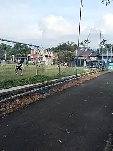 A short video shows a soccer field with multiple players dressed in jerseys of white and black with numbers. The camera is positioned outside the field's fence, capturing the action from a side view. A goal net is visible on the left side. Players are running, some chasing the ball. After a brief pause, the camera continues to record as the players continue to engage in the game. The weather appears clear. Buildings are visible in the background behind the field, as are trees. The ground visible in front of the fence is black asphalt.