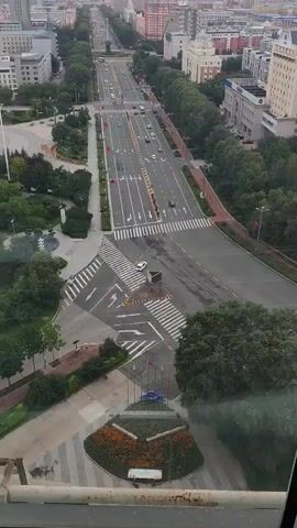 This is a high-angle aerial view of a wide, multi-lane city street lined with trees and buildings. The street has multiple lanes of traffic, pedestrian crosswalks, and what appear to be traffic islands. Cars are visible on the road. On the left side of the street, there is a park-like area with trees and a pathway. On the right side, there are more trees and buildings. In the distance, the city stretches out with more buildings under a cloudy sky. The video pans slightly to the right, showing more of the cityscape. There are no people prominently featured in the video. The overall environment appears to be urban.