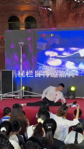 A male performer is on a red carpeted stage, performing a split. He is wearing a white jacket over a white t-shirt and black pants. A large LED screen behind him displays a variety of images, including a number "1" in a red circle and abstract colorful graphics. A crowd of people, mostly facing away from the camera, are watching the performance. Some are holding up phones to record. The environment appears to be outdoors, with a brick building visible in the background and decorative lights strung overhead.