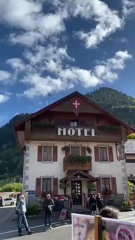 The video pans across a charming Swiss village on a bright, sunny day with a few puffy clouds in the blue sky. The focus is on the traditional architecture, with alpine-style buildings featuring wooden balconies adorned with flower boxes. A hotel is prominently displayed, marked with a large "HOTEL" sign and the Swiss flag on its roof. People are seen walking around the village, dressed in casual clothing. In the background, lush green mountains rise, completing the picturesque alpine setting. A Scott banner can also be seen on one of the buildings.
