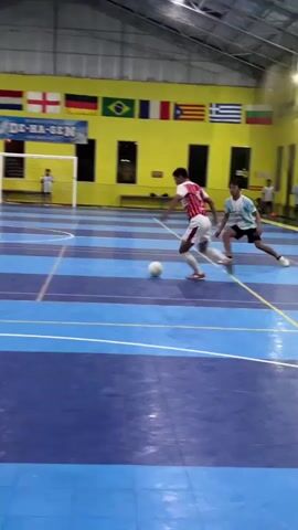 An indoor soccer game is in progress on a blue and purple court. A player in a white and red jersey with the number 17 on the back, identified as F. OZAN, is in the foreground, facing away from the camera. Other players are visible on the court, with one player in a white and blue jersey attempting to control a soccer ball. In the background, a yellow wall displays flags of various countries, including Mexico, Spain, Czech Republic, Portugal, England, and Brazil. A goal net is positioned at the far end of the court. The environment appears to be a sports facility with a high ceiling and overhead lighting.