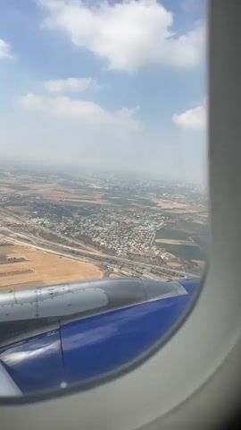 A view from an airplane window shows a landscape below with buildings, fields, and a highway. The wing of the airplane, painted blue and silver, is visible in the foreground. The sky is blue with scattered white clouds. The image is stable, suggesting the plane is in flight.