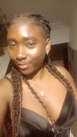 A woman is taking a selfie video. She has dark skin and is wearing a black corset, gold necklace and earrings, and a nose ring. Her hair is styled in long braids. She moves around and poses while smiling at the camera. The background changes, including a staircase, red wall, and a poster for "M-TRENDS." Music is audible in the background.