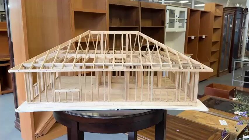A model of a house under construction is displayed on a dark round table. The model is made of wood and shows the framing of the walls and roof, with the interior floor plan visible. In the background, there are large wooden bookshelves filled with various items, suggesting the setting is a store or warehouse. The lighting is bright and even. The camera pans slowly around the model, offering different angles of the construction details. No people are visible in the immediate vicinity of the model.