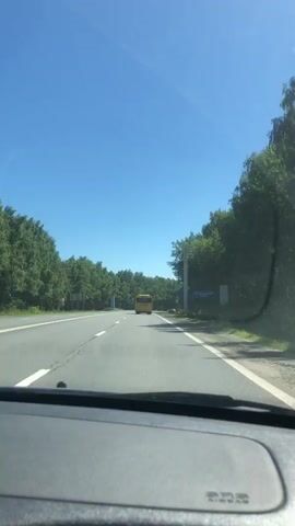 The video shows a forward-facing view from inside a vehicle traveling on a road. A yellow bus is visible in the distance, ahead of the vehicle. The road is paved with multiple lanes, and bordered by lush green trees on both sides. The sky is a clear, bright blue, indicating sunny weather. There are signs visible on the right side of the road. The interior of the vehicle, specifically the dashboard, is partially visible in the foreground. The camera appears to be mounted on the dashboard or windshield. The vehicle is in motion, as evidenced by the slight blur and the perspective shift relative to the stationary objects.