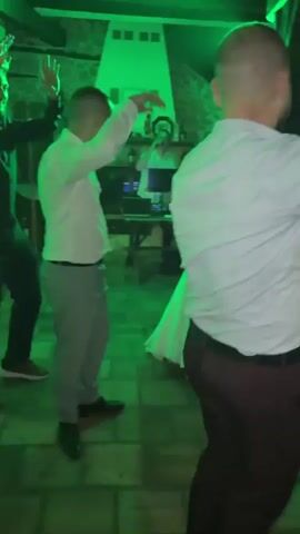 A wedding party is underway. A bride in a white dress dances joyfully while guests in formal attire, mostly men in white shirts, dance around her. A DJ booth is visible in the background. The atmosphere is celebratory, with lights and music creating a festive mood.