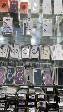 This media file showcases a retail display of mobile phone accessories in Bireuenmeunasahreuleut, Indonesia. The scene is an indoor setting, likely a mobile phone shop or a stall within a market, with shelves filled with various items.

The primary subjects are **objects**, specifically mobile phone cases and chargers. There are numerous phone cases displayed, many with magnetic charging rings, indicating compatibility with MagSafe technology. These cases come in a variety of colors, including white, purple, gold, and black. Several boxes of phone chargers are also visible on the top shelf, labeled with brands and wattage.

The **scene** is a close-up view of shelves stocked with these accessories. The lighting suggests it is daytime, though the indoor setting makes it difficult to determine the exact time of day or weather conditions. The presence of retail packaging and the organized display suggest a commercial environment.

**Notable details** include the brand names on some of the phone boxes, such as "Realme C85," "Vivo Y11d," and "OPPO A6x." Some of the phone cases have text indicating their model compatibility, for example, "For SM A25 5G Magnetic" and "A54 5G." On the bottom shelf, there are boxes with handwritten labels, likely indicating stock or pricing, such as "FOR SA A17 BLACK" and "OP A55." A small bottle of clear nail polish is also placed among the phone cases on the second shelf from the top, an unusual item in this context. A blurred figure of a person, possibly a shopkeeper or customer, can be seen behind the second shelf of phone cases, adding a human element to the scene. There are no overt expressions of emotion, as the focus is on the products.

The **location context** of Bireuenmeunasahreuleut, Indonesia, is provided. This suggests the items and the retail environment are specific to this region.