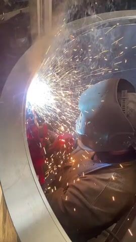 The video shows a person welding inside a large metal ring. The welder wears a protective mask, brown work jacket, and red gloves. Bright sparks shower from the welding point, partially obscuring the view. The ring appears to be made of stainless steel, and the environment around the welder is an industrial setting, with glimpses of other metal structures and equipment visible. The welding activity is the central focus.