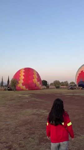 The video shows a field where hot air balloons are being prepared for flight. The background is a clear, light blue sky at dawn or dusk. In the initial shot, a young woman with long dark hair, wearing a red jacket and jeans, walks toward two deflated hot air balloons. The one on the left is a patchwork of red, yellow, and white squares, while the one on the right has sections of red, white, blue, and gray. As she approaches, the camera pans to include more of the second balloon, which appears to be partially inflated. Two other individuals, a man with glasses wearing a beige top and a woman in a black coat, stand beside her, watching the preparations. Several vehicles, including a black pickup truck, are parked nearby. Workers in safety vests and red jackets are around the partially inflated balloon, appearing to be making final adjustments to the basket. The balloon's top section has the word "Volare" and a website address printed on it. The ground appears to be a mix of soil and sparse grass.