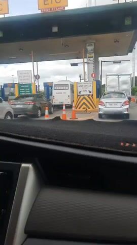 The video shows a view from inside a car at a toll booth. Cars are lined up in lanes with orange traffic cones. There are signs on poles and overhead structures. One sign reads "NO VALID ETC DEVICE NO ENTRY". Another sign says "AVOID TAILGATING NEXT VEHICLE STOP HERE". A white bus is visible in one lane. The dashboard of the car is also visible, with a touchscreen display and buttons. The overall environment appears to be an outdoor toll plaza.