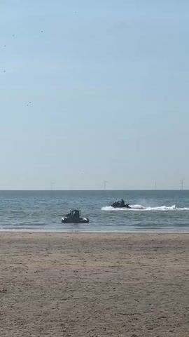 A wide shot of a beach shows a calm ocean with a clear blue sky. In the distance, several wind turbines are visible on the horizon. On the water, two jet skis are being ridden. One jet ski is closer to the shore and is moving slowly. The other jet ski is further out in the water and is creating a spray as it moves at a faster pace. There are a few people in the water near the shore, appearing to be swimming or wading. The sand of the beach occupies the foreground of the image.