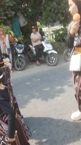 The video shows a parade or procession of people, including children, dressed in traditional clothing. The clothing appears to be black with gold and patterned accents, including headwear. Some individuals are wearing hijabs. The group is walking down a paved road, with trees and buildings visible in the background. There are parked motorcycles and other vehicles along the side of the road. Spectators can be seen watching the procession. The overall impression is of a cultural event or celebration.