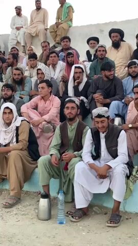 The video shows a group of men of various ages sitting in tiered seating, resembling bleachers. The men are dressed in traditional clothing, including turbans, shalwar kameez, and vests. Most of the men appear to be observing something out of frame. In the foreground, there are two water bottles and a thermos. The ground appears to be dirt or gravel. The lighting suggests it is daytime, possibly late afternoon or early morning.