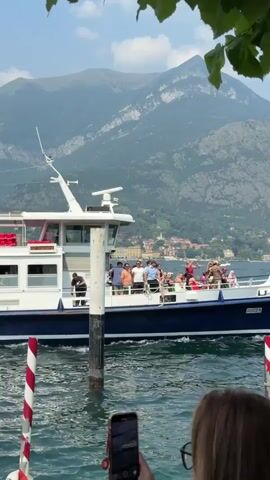 A ferry boat with passengers moves across a body of water. A person in the foreground is filming the ferry with a mobile phone. Mountains are visible in the background.