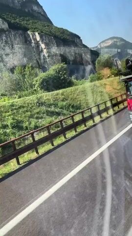 The video is a first-person perspective from a moving vehicle, likely a car or bus, traveling on a highway. The view is through a window that reflects the interior of the vehicle. Outside the window, a road with a white lane marker is visible, bordered by a metal guardrail. Beyond the guardrail, there is a grassy embankment and then a steep, rocky mountain face with sparse vegetation. The sky is clear and blue. A red truck is visible on the road ahead, moving in the same direction as the vehicle. The overall environment appears to be mountainous and rural, with a bright, sunny day. The camera is panning slightly as the vehicle moves, creating a sense of motion. There are no people clearly visible.