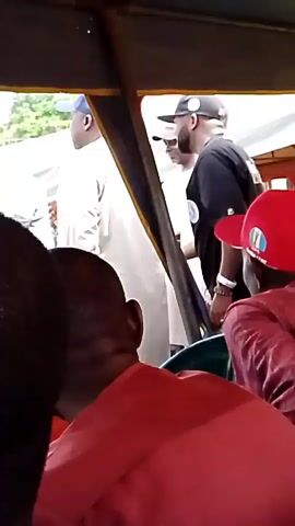 The video shows the interior of a vehicle, possibly a bus or van, with several people visible. In the foreground, the back of a person wearing a red shirt is visible. To their left, a person in a white outfit and a blue cap is seen from the side. In the middle ground, a person wearing a black t-shirt and a black baseball cap is facing away from the camera and looking to the right. To their left, another person wearing sunglasses and a white t-shirt is partially visible. Further to the right, a person wearing a red baseball cap is partially visible. The environment appears to be outdoors, with greenery and a white structure visible through the windows. The lighting suggests it is daytime.