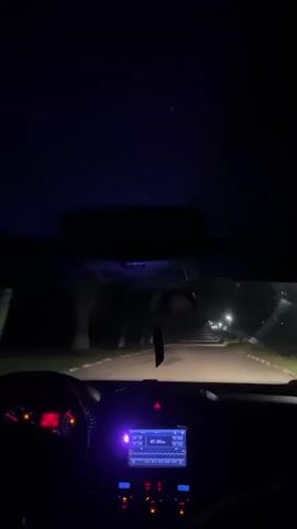 The video shows a view from inside a car driving down a dark road at night. The dashboard is visible, with the radio on. Streetlights line the road. The car is moving forward. The steering wheel can be seen, suggesting someone is driving. The overall environment is dark and quiet.