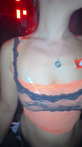 A woman is dancing at a club with red lighting. She is wearing a black and orange lingerie-style top with a silver necklace. She has brightly colored nails, several rings on her fingers, and a "HOT" sticker on her chest. She is smiling and laughing.