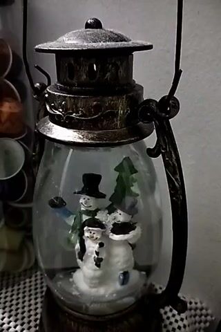 The video shows a close-up of a snow globe lantern. The lantern has a dark metal finish and a glass globe. Inside the globe, there are three snowmen and a Christmas tree. The snow globe appears to be sitting on a checkered surface. To the left of the lantern, a blurred cylindrical container filled with what appears to be coffee pods is visible. The background is a plain wall. The lighting is dim, suggesting the video was taken indoors. There is no discernible action or interaction occurring in the video. The snow globe itself is stationary.