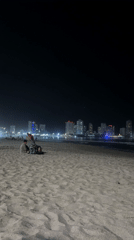 Two people sit on beach at night near city