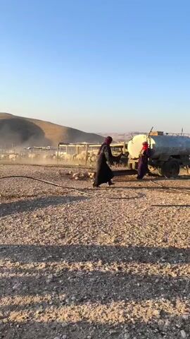 TWO PEOPLE, ONE WEARING A BLACK ROBE AND HEADSCARF AND THE OTHER A PURPLE ROBE AND RED HEAD COVERING, ARE STANDING NEAR A WATER TANKER TRUCK IN A DUSTY, OPEN AREA. IN THE BACKGROUND, A FLOCK OF SHEEP IS HOUSED IN A CORRAL. A BLACK GOAT IS NEAR THE WATER TRUCK. THE SUN IS SHINING, CASTING LONG SHADOWS, AND THE SKY IS CLEAR BLUE. THE ENVIRONMENT APPEARS TO BE RURAL OR SEMI-ARID.