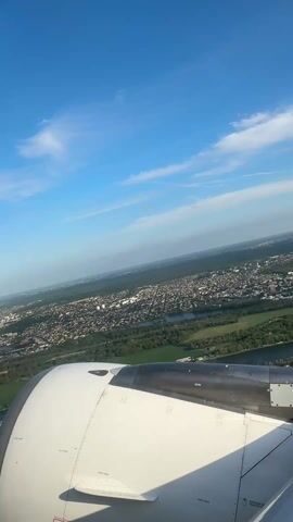 The view from an airplane window shows the landscape below and part of the plane's engine. The sky is blue with scattered white clouds.  Below, a dense cityscape extends toward the horizon. Patches of green, likely parks or fields, intersperse the urban area, and a wide river meanders through the landscape. The plane's engine is mostly white with some gray and black components visible.
