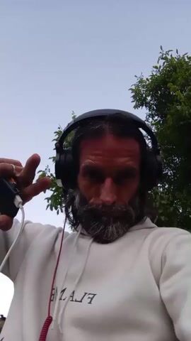 A man wearing headphones and a white hoodie, with a beard, is looking at the camera outdoors. He is holding up his left hand, with his fingers slightly spread. The man's face is red, and his eyes are dark. He is smiling. Trees are visible in the background.