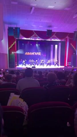 An elevated, static, wide shot from the perspective of an audience member in a theater captures a stage performance. In the foreground, the backs of numerous audience members are visible, seated in dark red plush theater seats. A bouquet of white flowers rests on one of the seats in the bottom left. On the stage, approximately twelve dancers, dressed in dark attire, are performing a choreographed routine. They move in unison, bending and gesturing. Behind them, a large screen displays the word "АВАНГАРД" (AVANGARD) in orange and purple text, with purple light streaks across the black background. The stage is brightly lit with purple and white lights, and red curtains frame the sides of the stage. The theater ceiling is also illuminated with purple lighting. Some audience members are holding up their phones to record the performance.