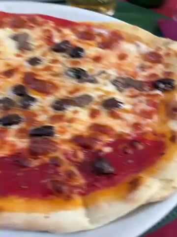 A close-up shot showcases a pizza, its surface featuring a vibrant mix of tomato sauce, melted cheese, and scattered black olives. The crust, golden and slightly charred, suggests a well-baked base. The pizza is presented on a white plate, with glimpses of other tableware and a green surface hinting at a restaurant or dining setting.