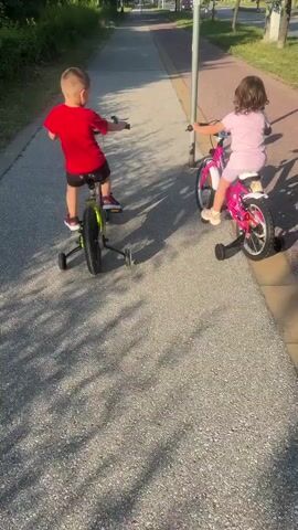 Two young children are riding bicycles with training wheels on a paved path. The child on the left is wearing a red shirt and black shorts. The child on the right is wearing a pink dress and white shoes. The path is lined with grass and trees. The sun is shining, casting shadows on the path. The children appear to be moving forward, but their exact speed and direction are unclear. The video is shot from a low angle, looking up at the children. There is a pole with a light fixture on the right side of the path. The overall scene is one of childhood activity in an outdoor environment.