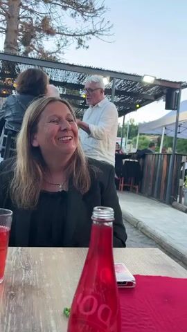 A woman with long blonde hair smiles and laughs while sitting at a table outdoors. She is wearing a black blazer over a black shirt. A red bottle of drink and a red napkin are on the table in front of her. A man in a white shirt stands behind her and to the right, looking down at something in his hands. In the background, other people are visible sitting at tables under an awning. The scene appears to be a restaurant or outdoor seating area.